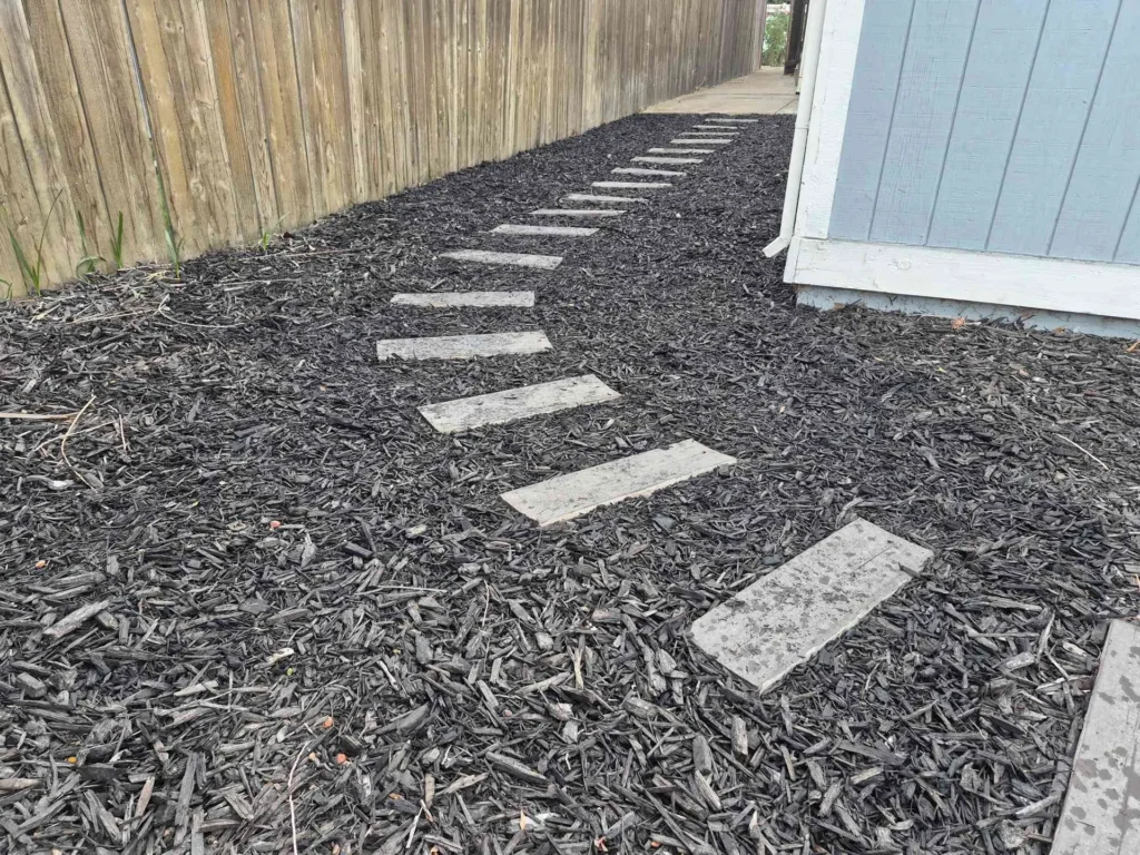 Rectangular stepping stones through dark mulch beside a wood fence and house.