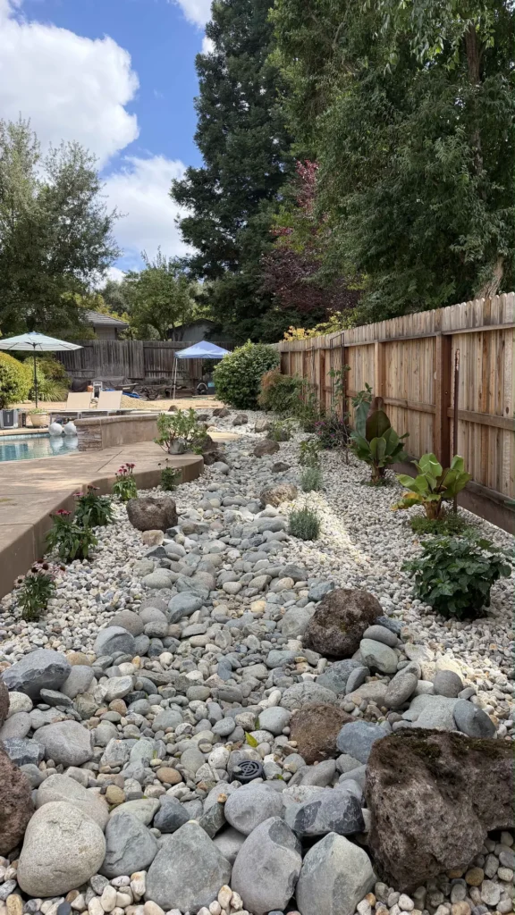 A rock-lined planting bed with boulders and flowering plants alongside a fence and pool deck.