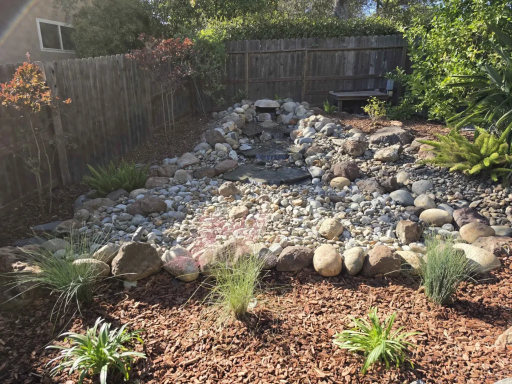 A small cascading water feature surrounded by river rocks, boulders and mulch.