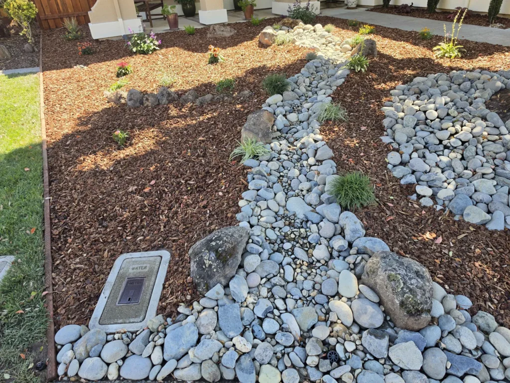 A winding river-rock bed through bark mulch with boulders and small flowering plants.